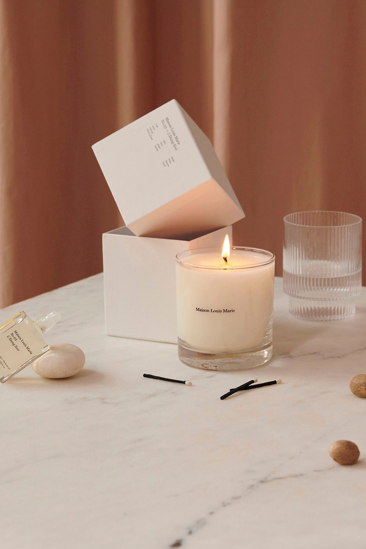 Poducts with the No.03 scent from maison louise marie. A scented candle and perfume oil standing on marble with a glass of water and ingredients in the scent.