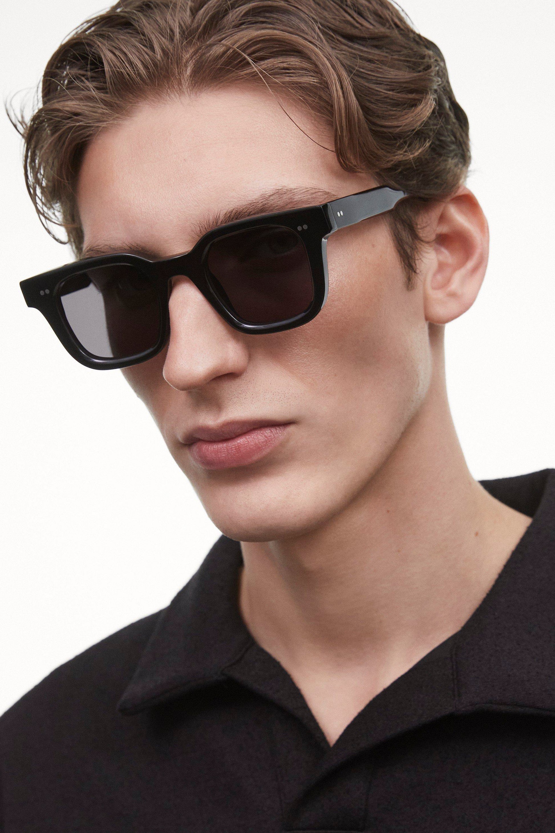 Closeup view of the Black Chimi 04 sunglasses on a male model. The glasses have square frames and a dark lens. 