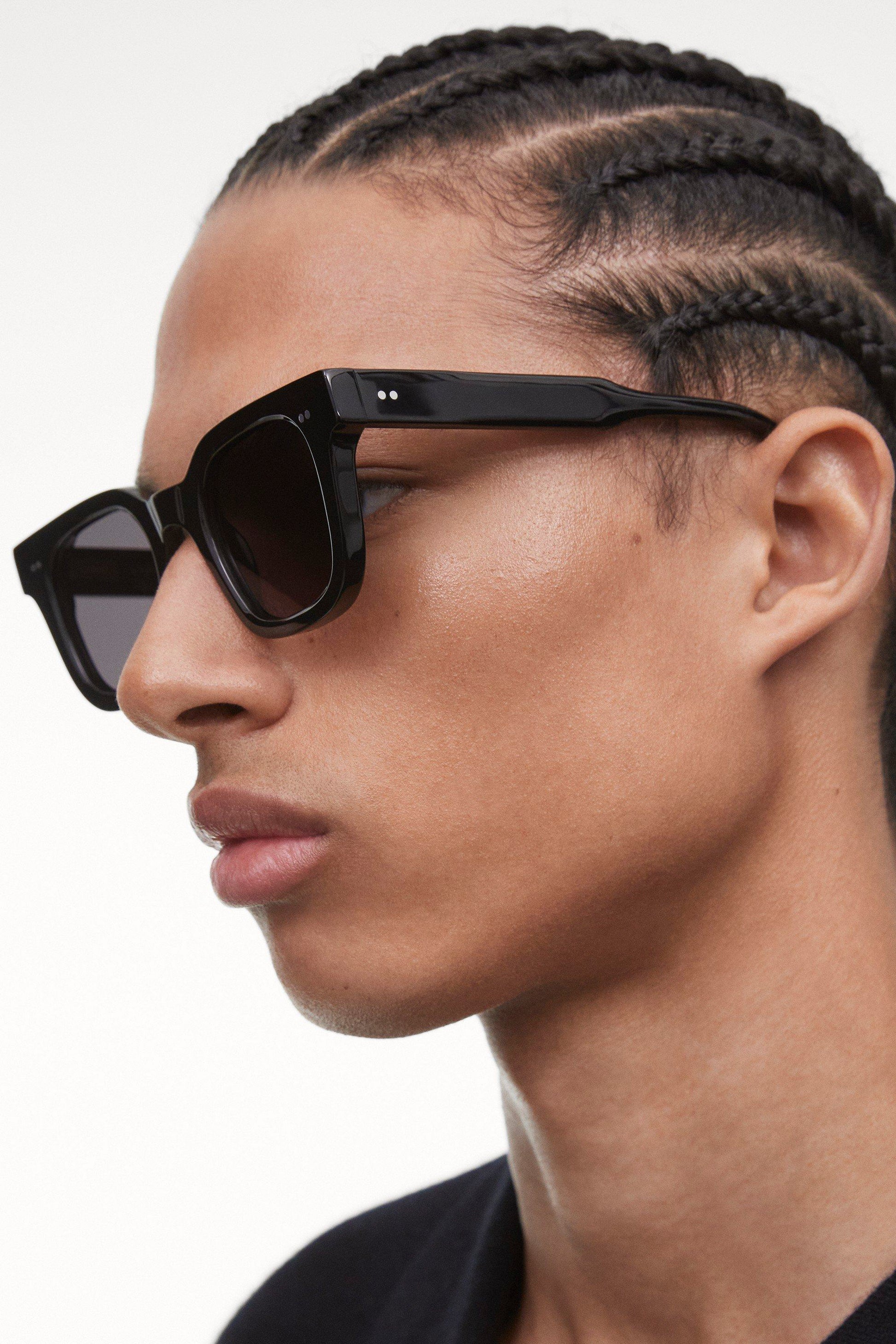 Side and closeup view of the Black Chimi 04 sunglasses on a male model of color with cornrows. The glasses have square frames and a dark lens. 