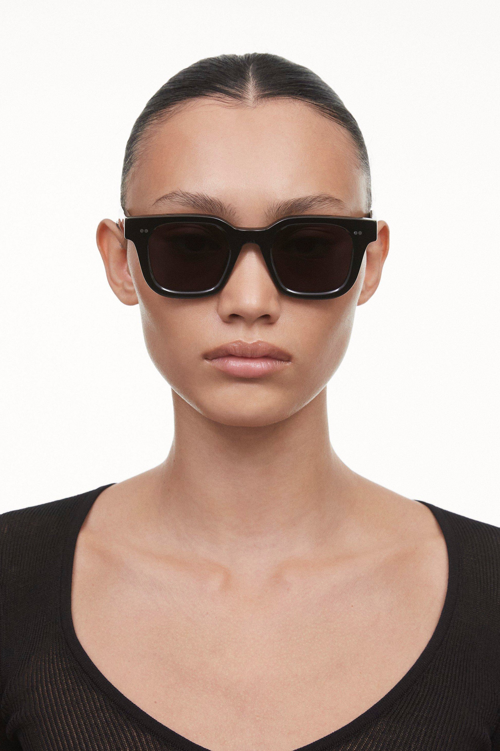 Black Chimi 04 sunglasses on a female model with brown hair. Featuring square frames and a dark lens. 
