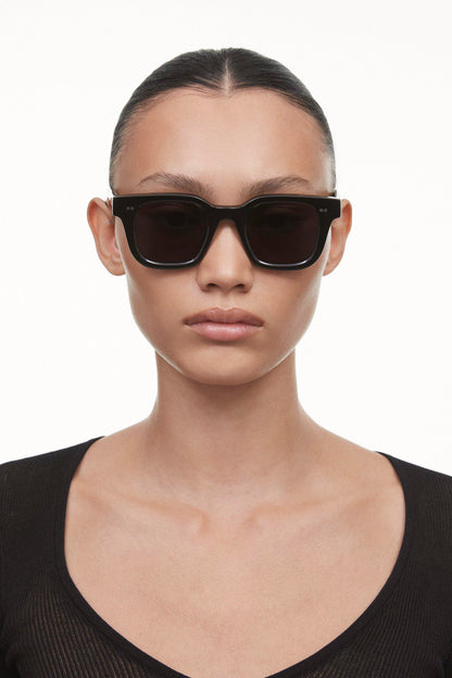 Black Chimi 04 sunglasses on a female model with brown hair. Featuring square frames and a dark lens. 