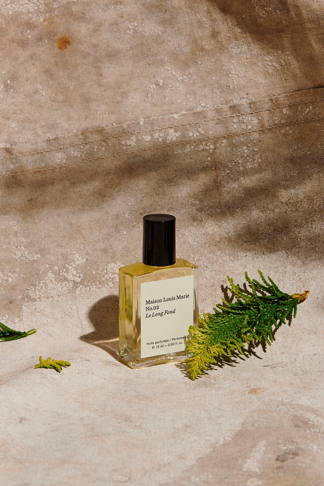 A bottle of Maison Louis Marie No. 02 Le Long Fond perfume oil placed with small green branches of foliage on textured fabric background. 