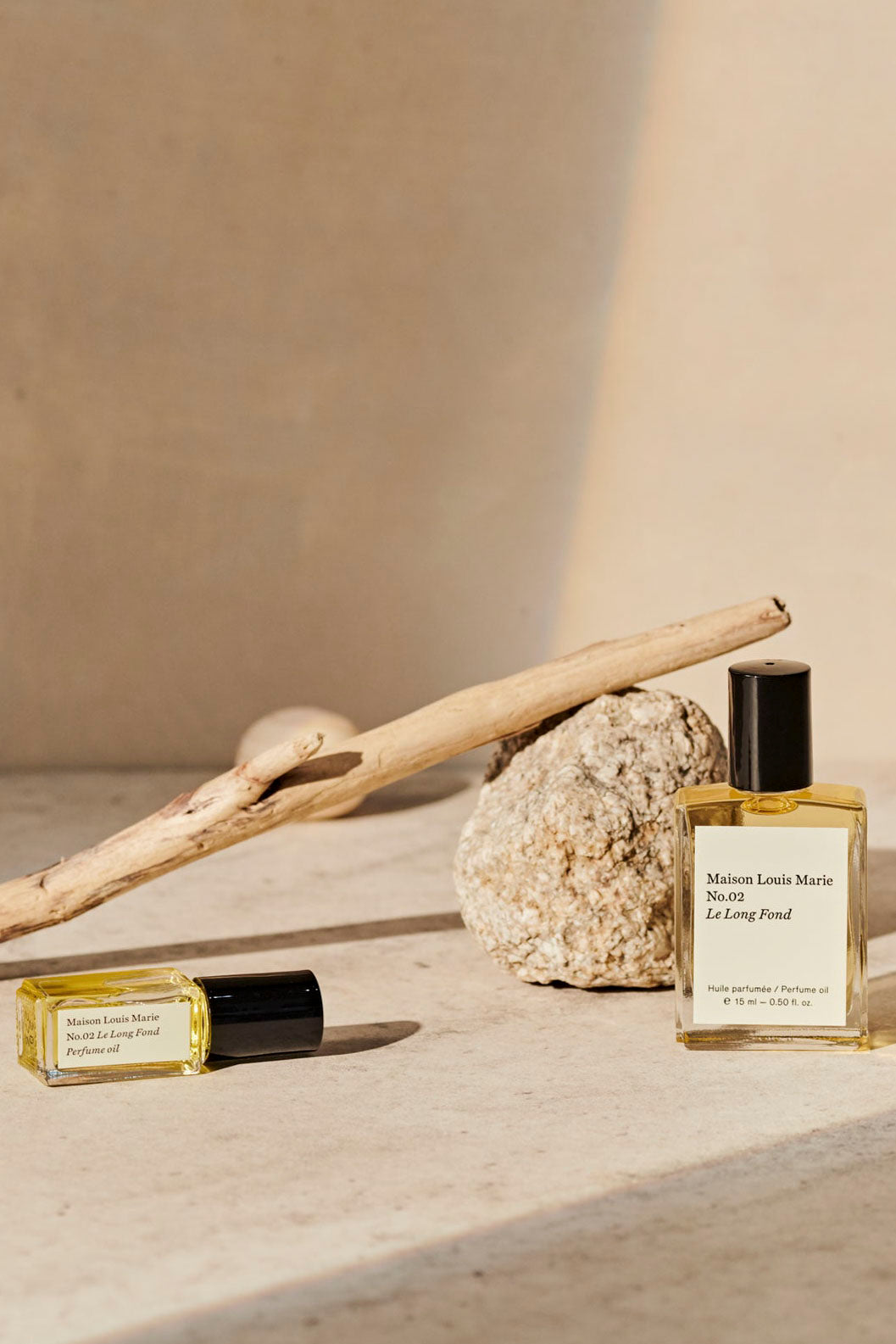 Two bottles of Maison Louis Marie No. 02 Le Long Fond perfume oil in an artistic set up with a treebranch and a stone. 