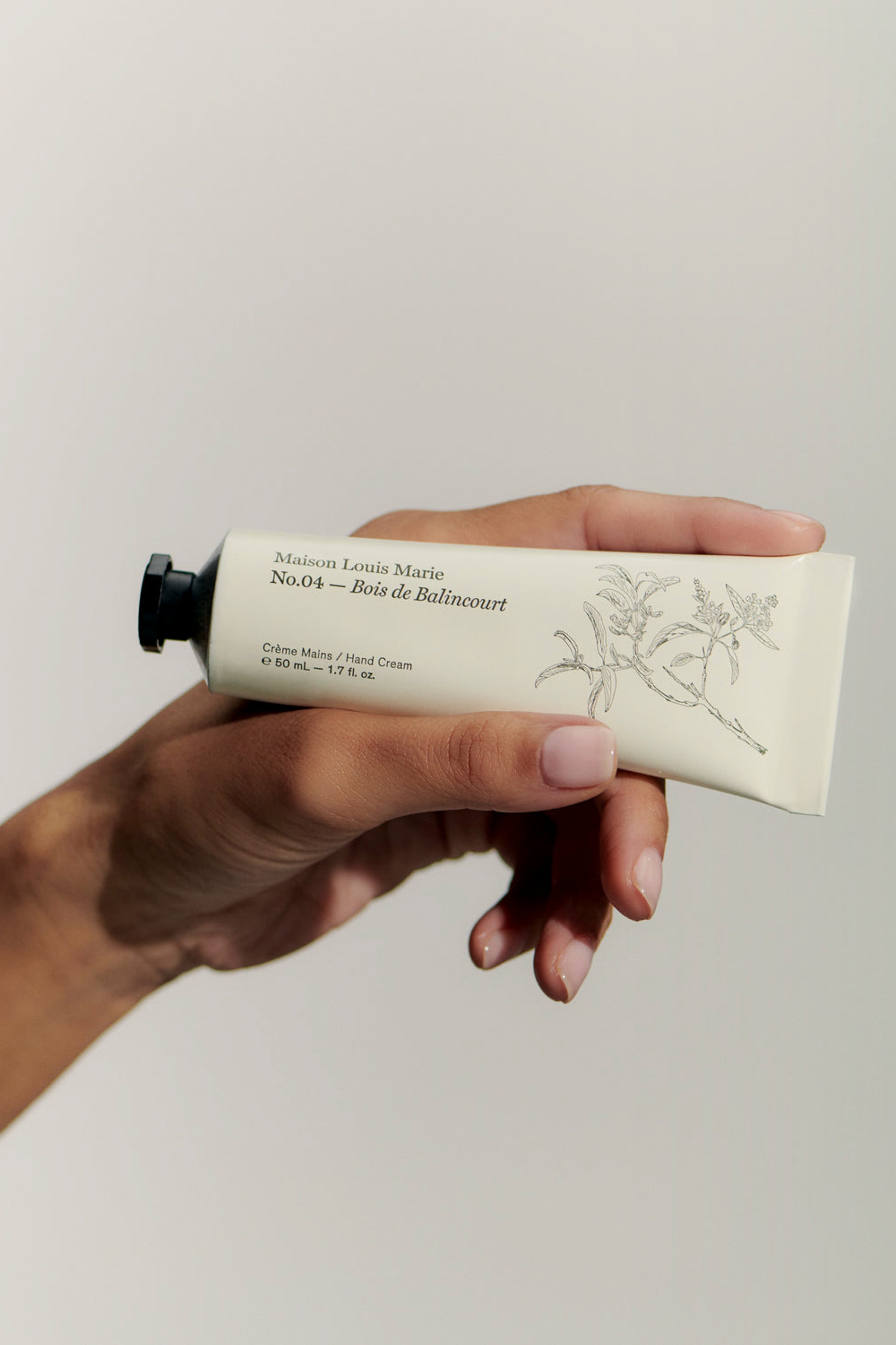 Hand holding a tube of handcream from Maison Louise Marie with No. 04 Bois De Balincourt smell. The tube is white with hand-drawn flowers and a black cap. 
