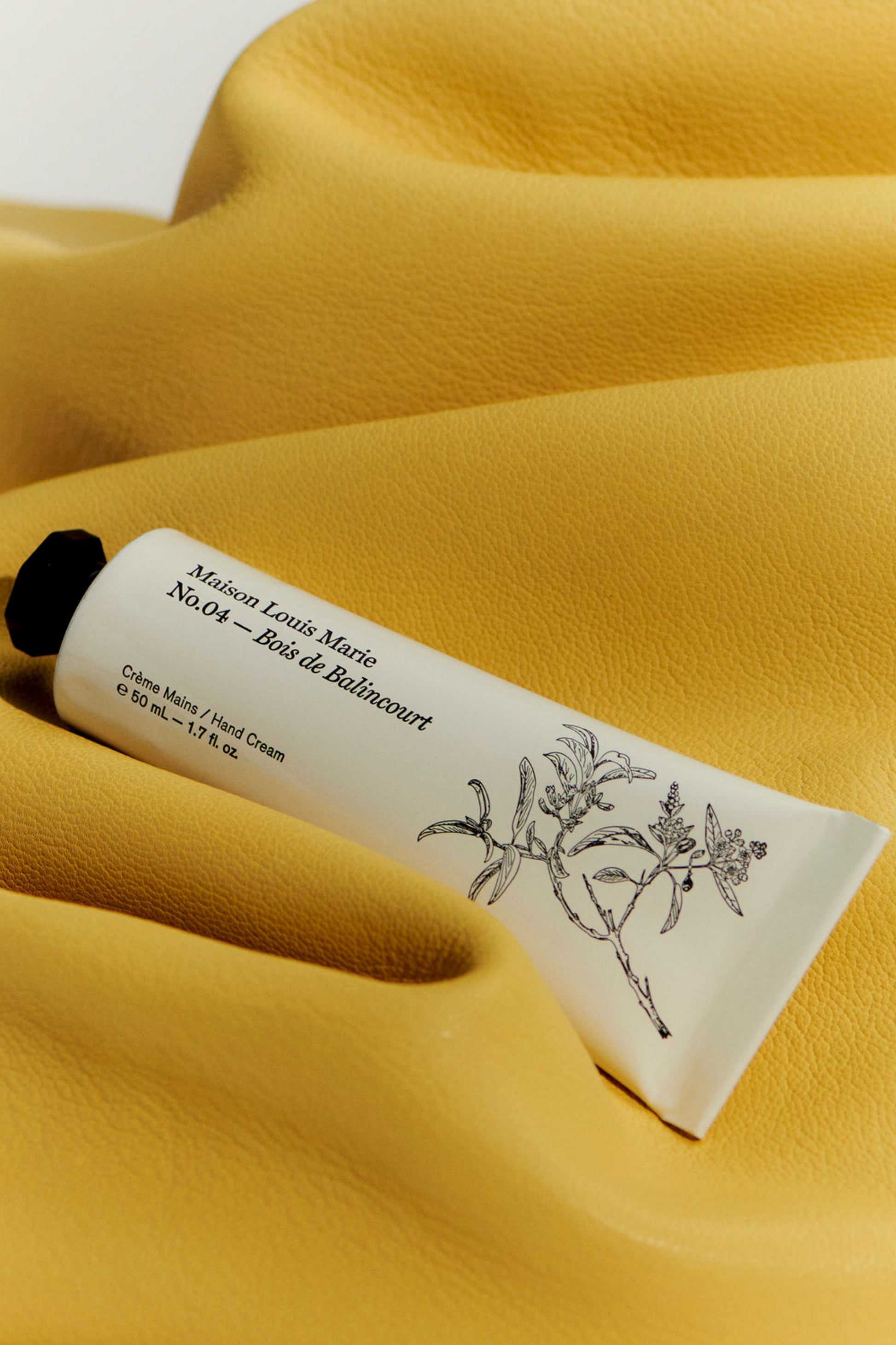 Handcream from Maison Louise Marie with No. 04 Bois De Balincourt smell. The tube is white with hand-drawn flowers and a black cap and is on a yellow velvet background. 