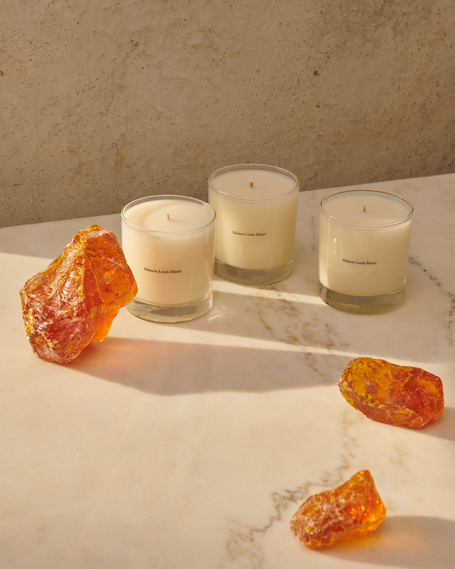 Three scented candles from maison louise marie. Standing on marble alongside orange stones. 